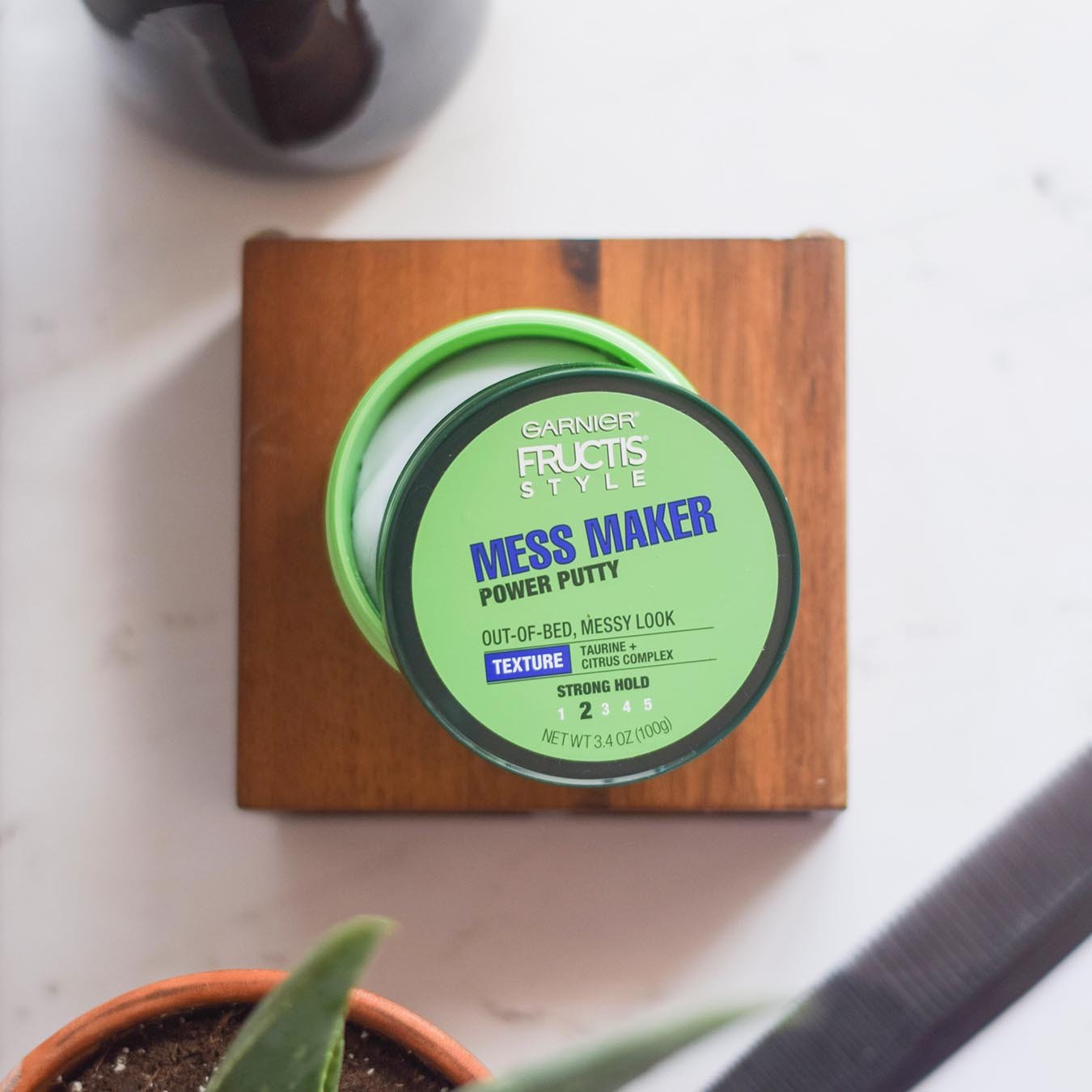 Garnier Fructis Style Mess Maker Power Putty ajar on a wooden block on white marble next to a potted aloe plant, black comb, and black vase.